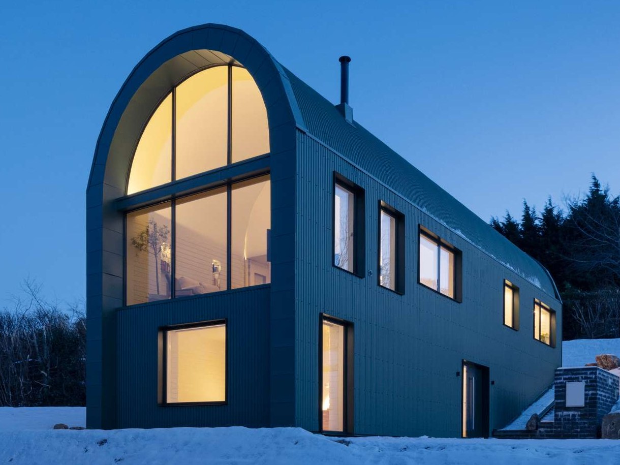 This modern home draws inspiration from the green corrugated steel sheds scattered across the Wicklow landscape, embracing their utilitarian grace while subtly elevating every detail. Its elongated form, curved roofline, and semi-submerged base follow the natural slope of the site, preserving its topographical character and minimizing excavation.