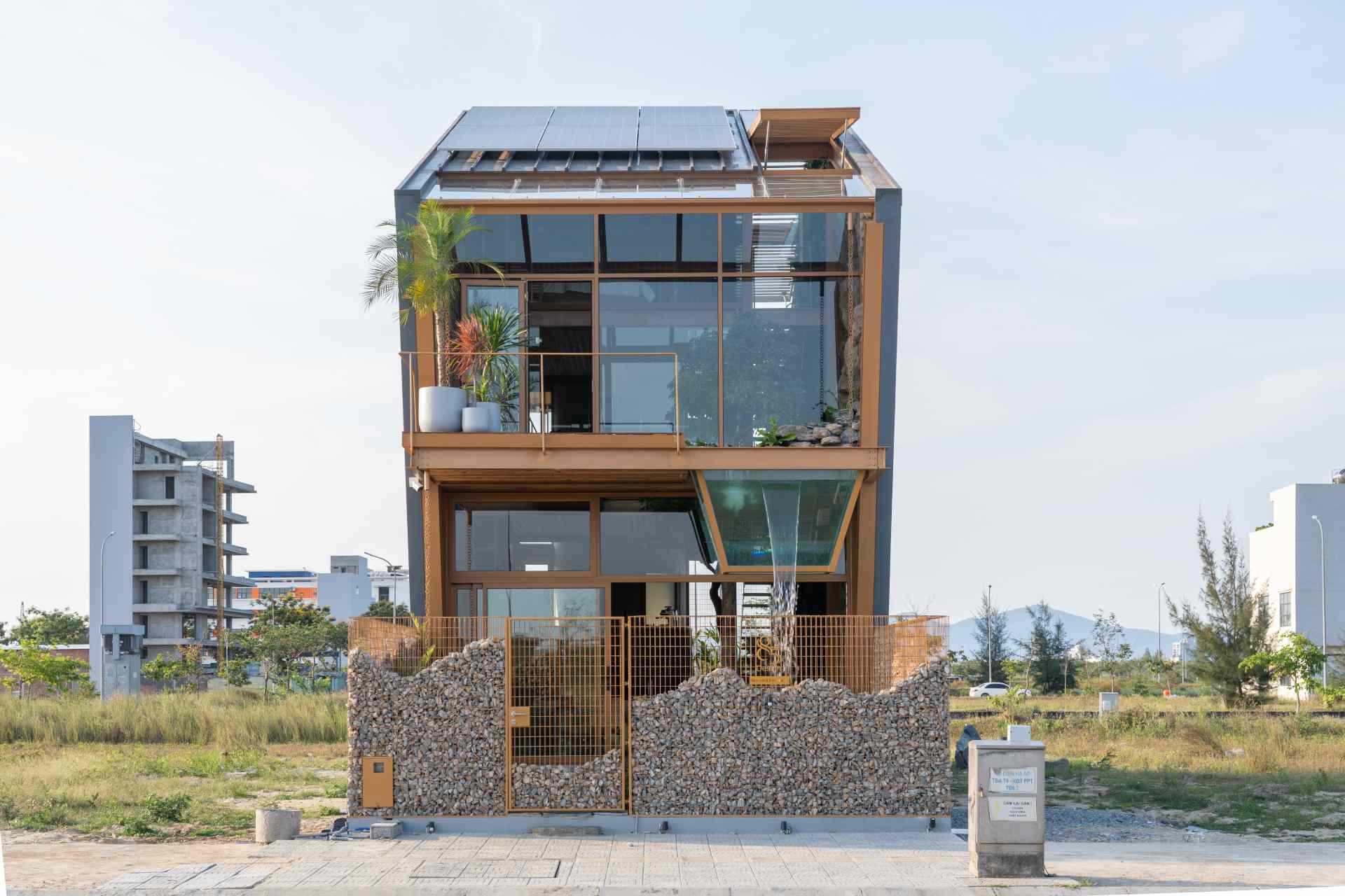 The exterior of this live-work building blends modern lines with natural materials, steel framing, warm wood, and wide glass panels. A glass box extending from the upper floor doubles as a water feature, while a stone-filled fence below adds texture and an earthy contrast.