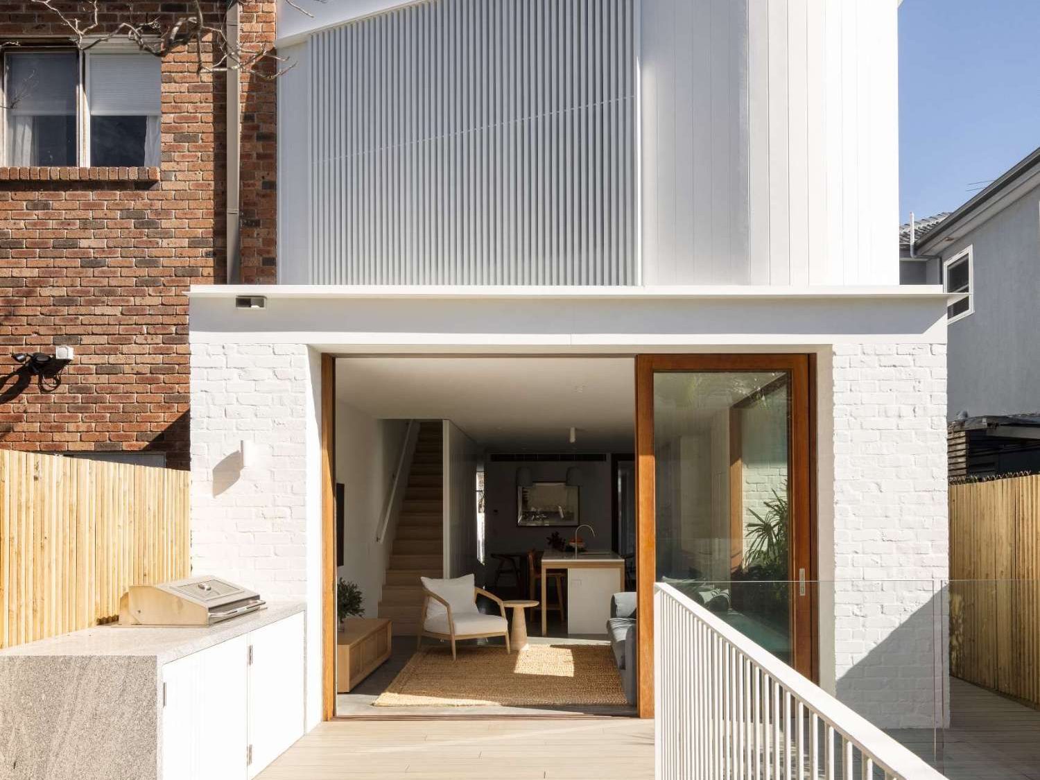 A crisp white volume with vertical battens above and painted brick below. Timber-framed glass doors slide open to reveal a light-filled living space that flows effortlessly to the deck, creating a breezy indoor-outdoor connection.