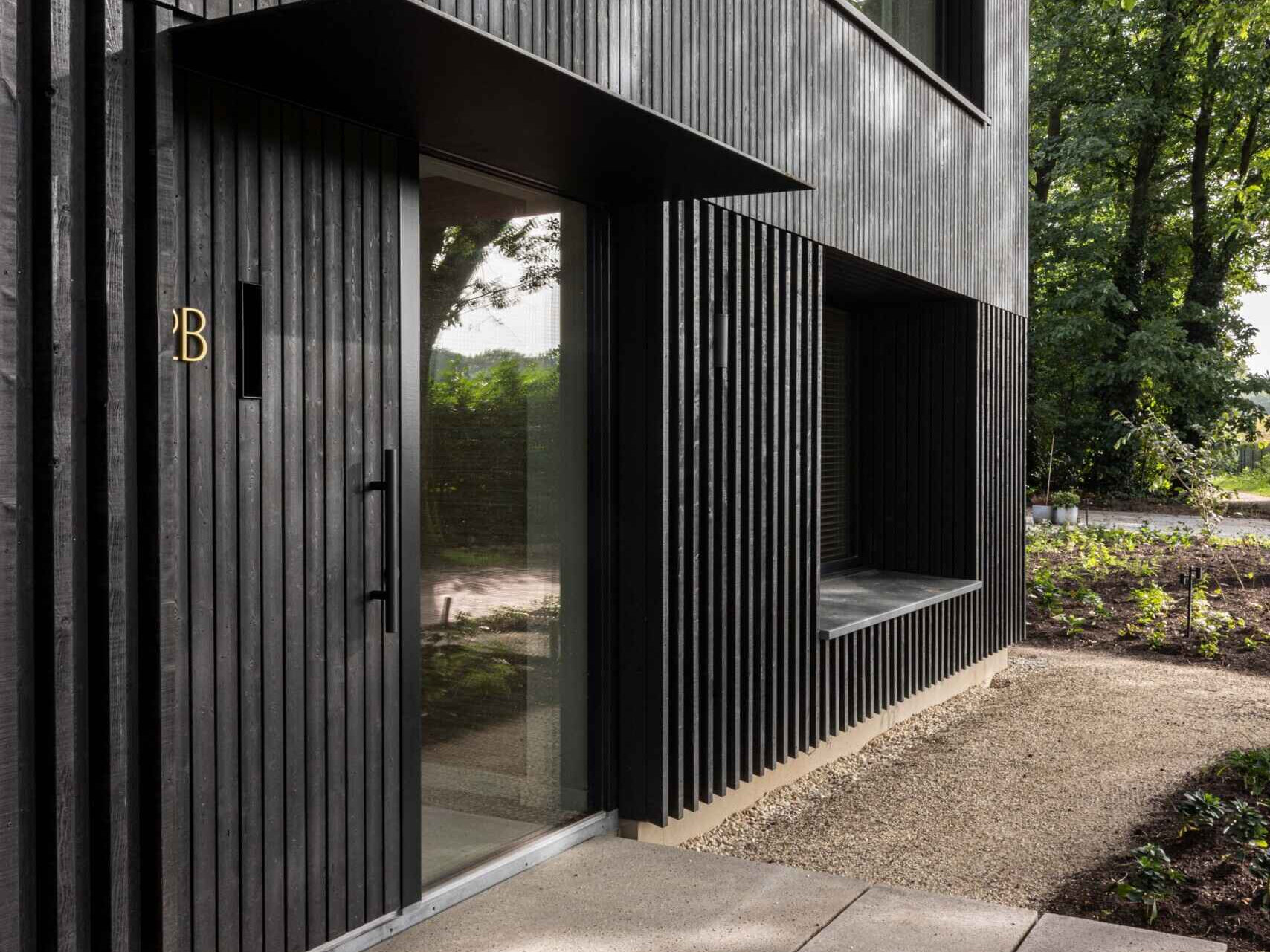 This striking modern home showcases the power of simplicity through its dark wood cladding and clean, rectangular form. Vertical wood slats add texture and rhythm to the facade, while the deep black finish helps the structure blend seamlessly into its natural surroundings. Generously sized windows are thoughtfully positioned to capture views of the landscape and invite daylight into the interior, all while maintaining the home’s strong architectural lines. 