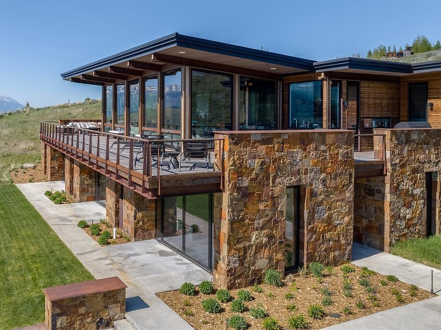 Nestled into the hillside like it naturally belongs there, this home embraces its mountain surroundings with a rugged elegance. The living roof, blanketed in grass, blends almost seamlessly into the wild landscape, while stacked stone walls ground the structure with earthy warmth. Generous windows invite the panoramic views inside, offering a daily dose of the snow-capped peaks beyond. It's the kind of place that feels both tucked away and wide open, perfect for quiet mornings or evenings that stretch out under the stars.