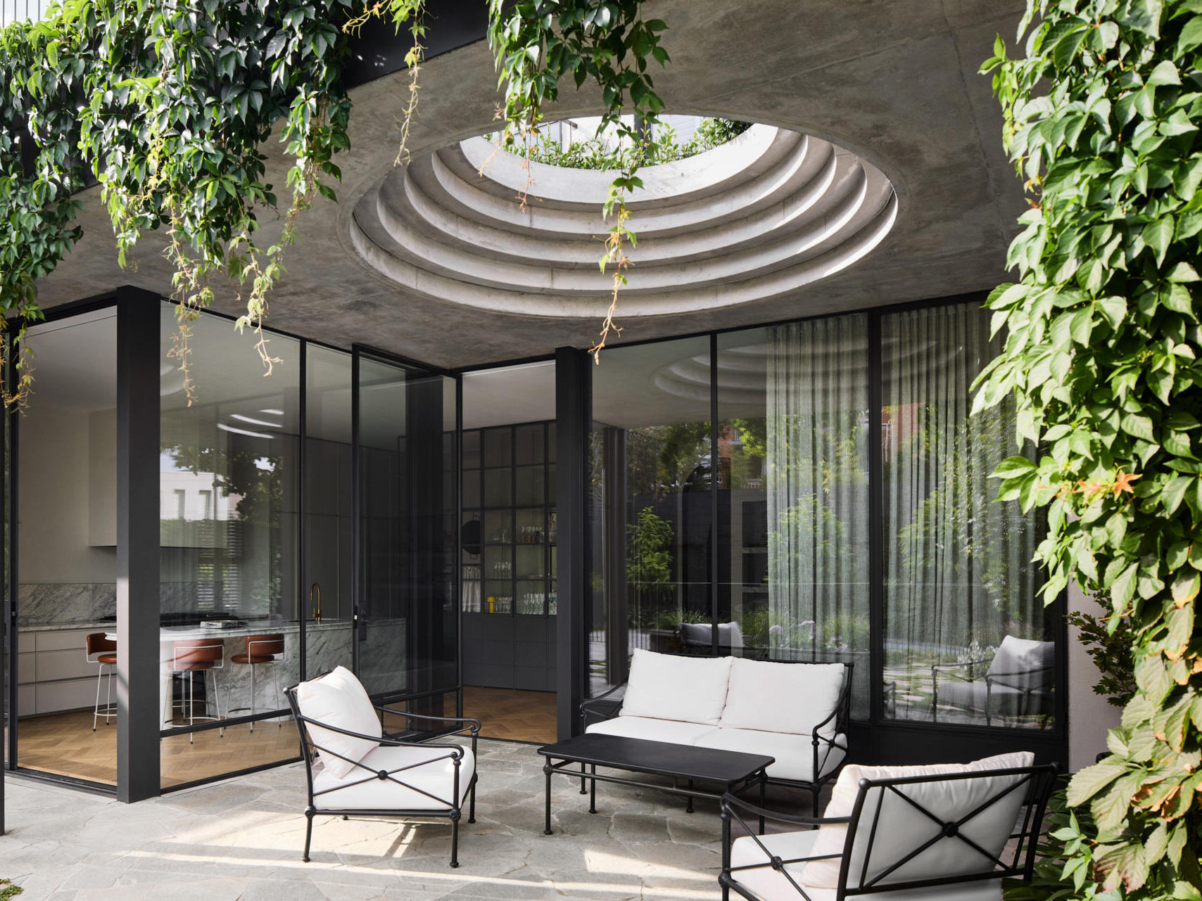 A defining feature of the rear of this remodeled home is a courtyard with a large circular cut-out overhead, a bold geometric move that invites sky views and dappled sunlight into the space below. Positioned above the outdoor dining area, the cut-out frames the moment, turning the patio into more than just a transitional space.