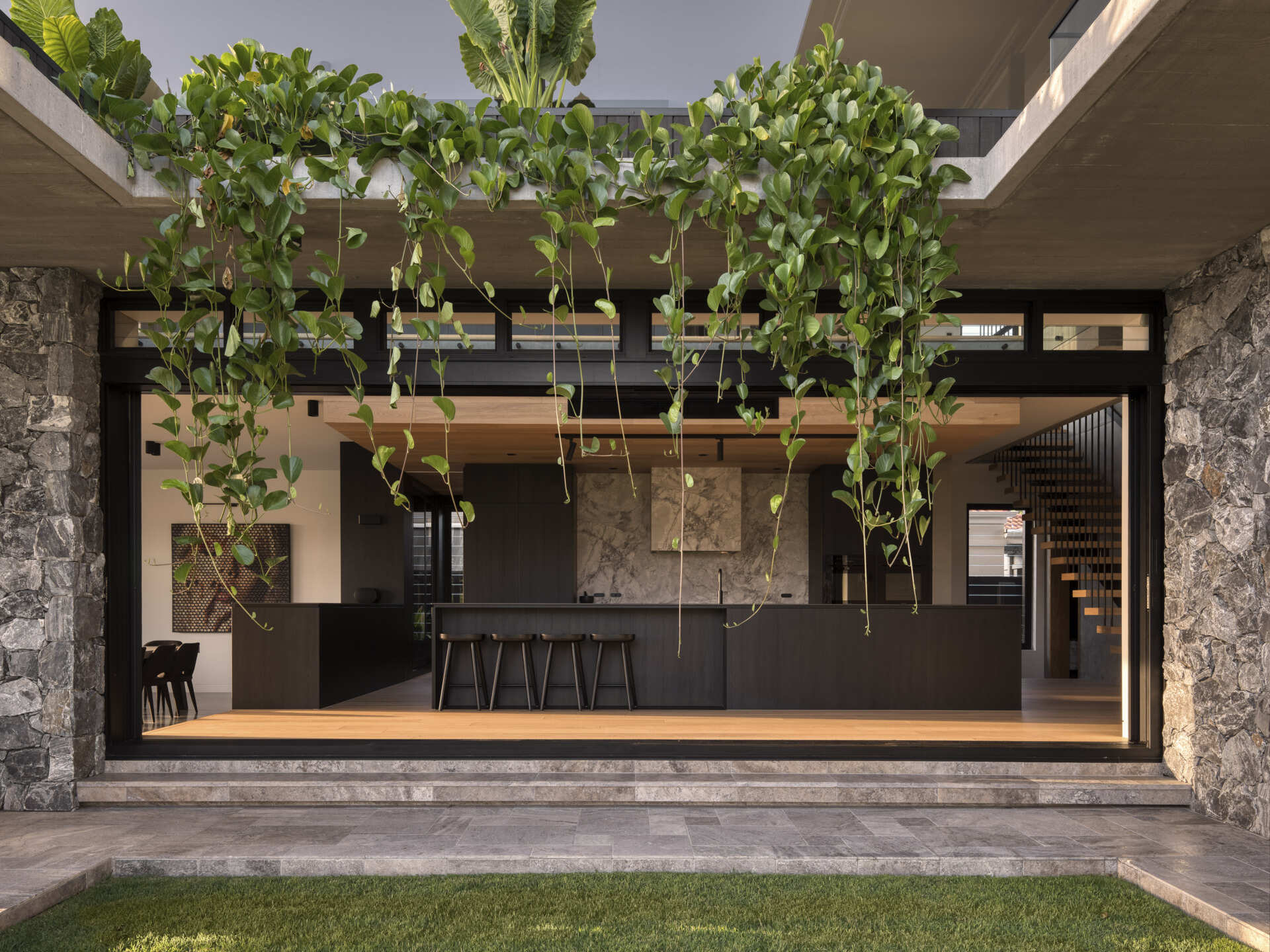 A modern black kitchen has sliding glass doors that connect it to the backyard, while greenery hangs down from the level above.