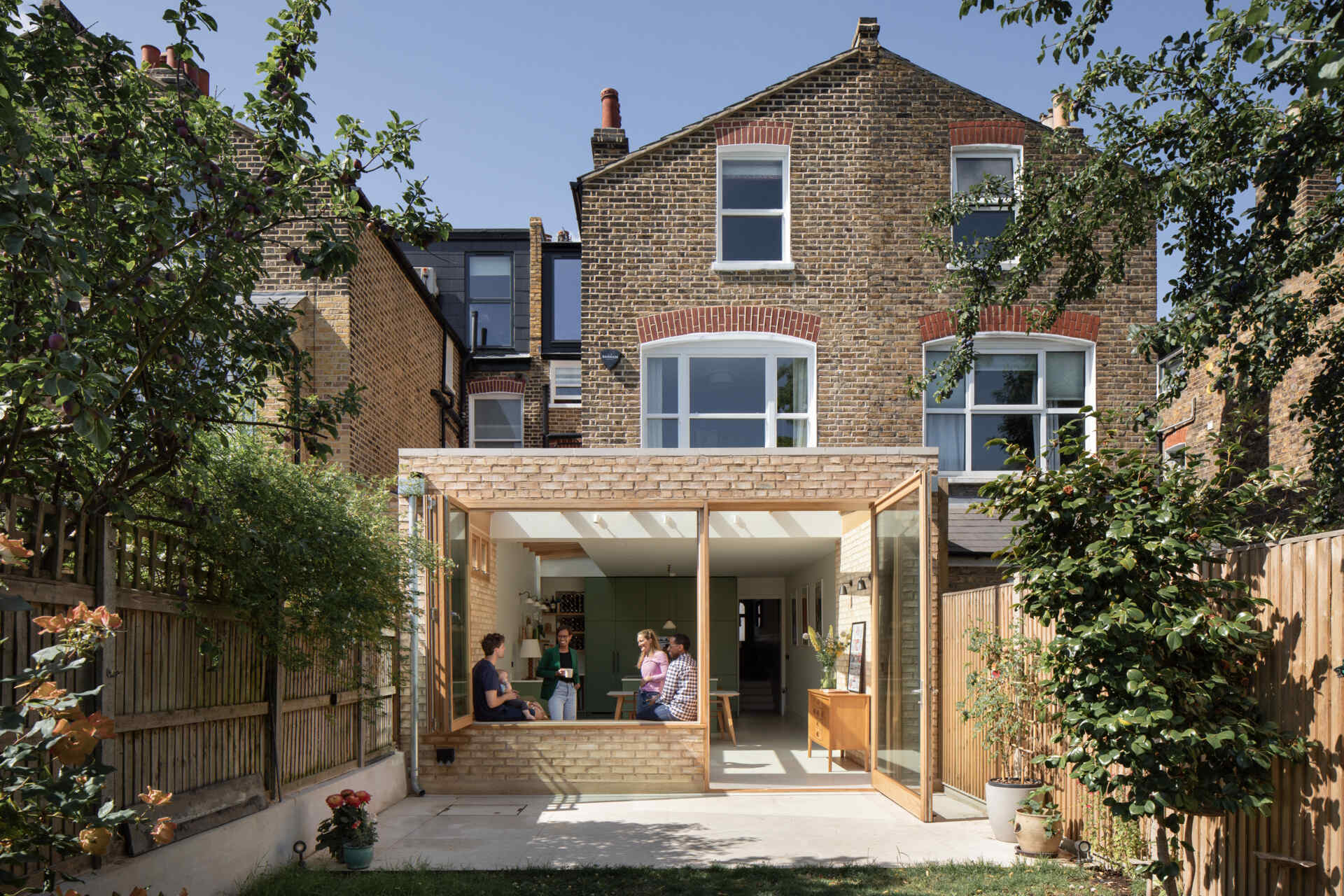 From the garden, the extension reads as a light-filled counterpoint to the home’s classic brick. Warm wood framing and generous glass panels open wide to the backyard, allowing sunshine to move freely across the stone floor and into the kitchen beyond.