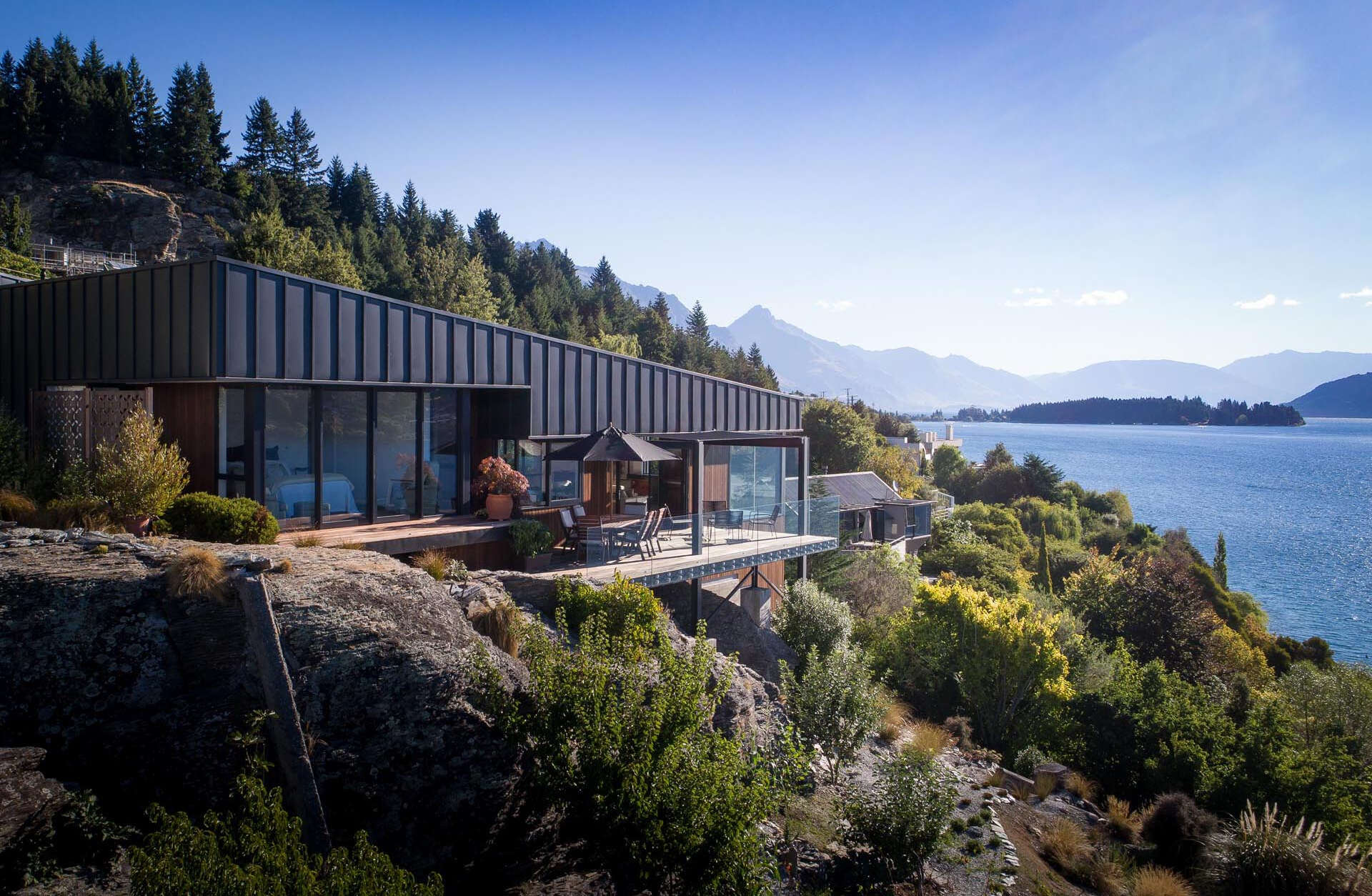 A modern home with two wedge shaped volumes clad in matte black metal and wood step down a steep Queenstown site while framing views of Lake Wakatipu.